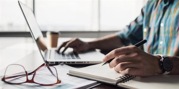A man writes on a notebook while typing on a laptop.