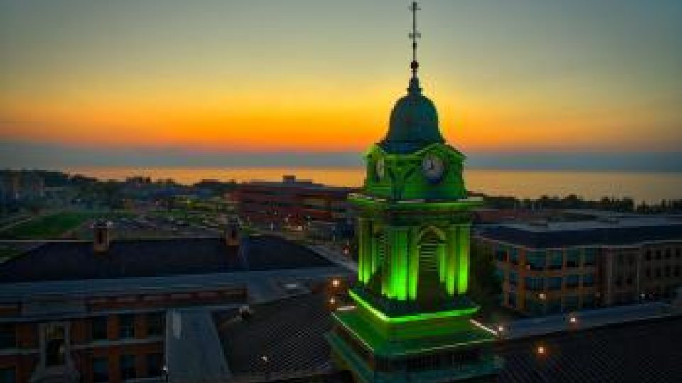 Aerial view of Sheldon Hall at sunset, with the building illuminated in green lights and the sun setting over a distant lake.