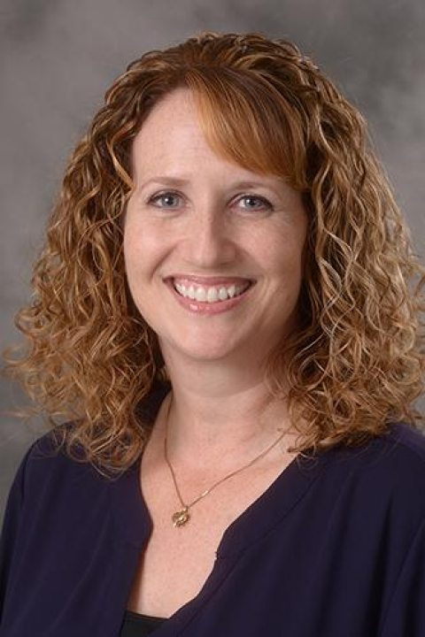 Portrait of Melissa Krass, a woman with shoulder-length curly red hair, wearing a dark purple top, smiling at the camera.