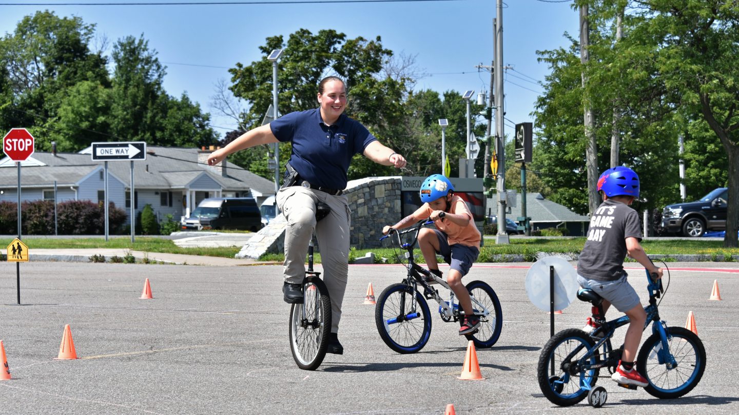 A University Police officer on a unicycle and young children on bicycles riding around an obstacle course