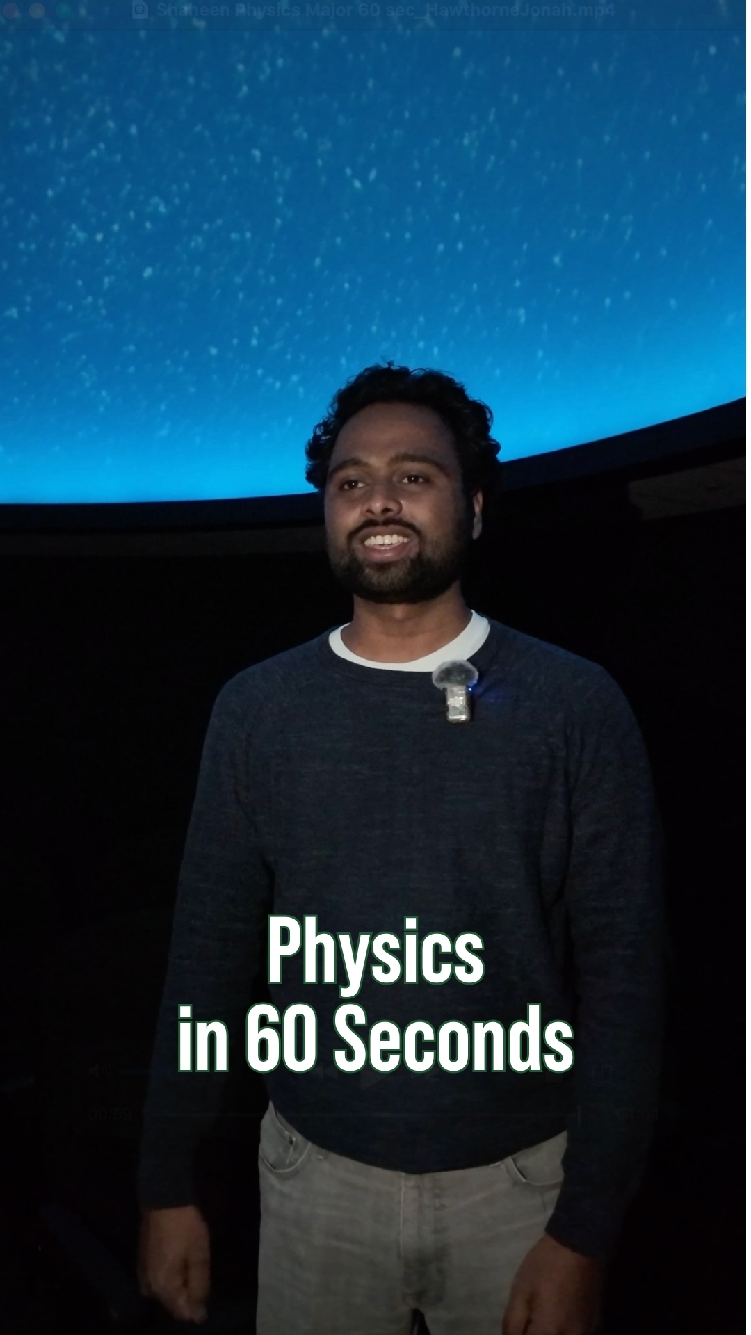 A student stands under a star-filled planetarium dome in a dark room with a microphone clipped to his shirt, with on-screen text reading “Physics in 60 Seconds.”