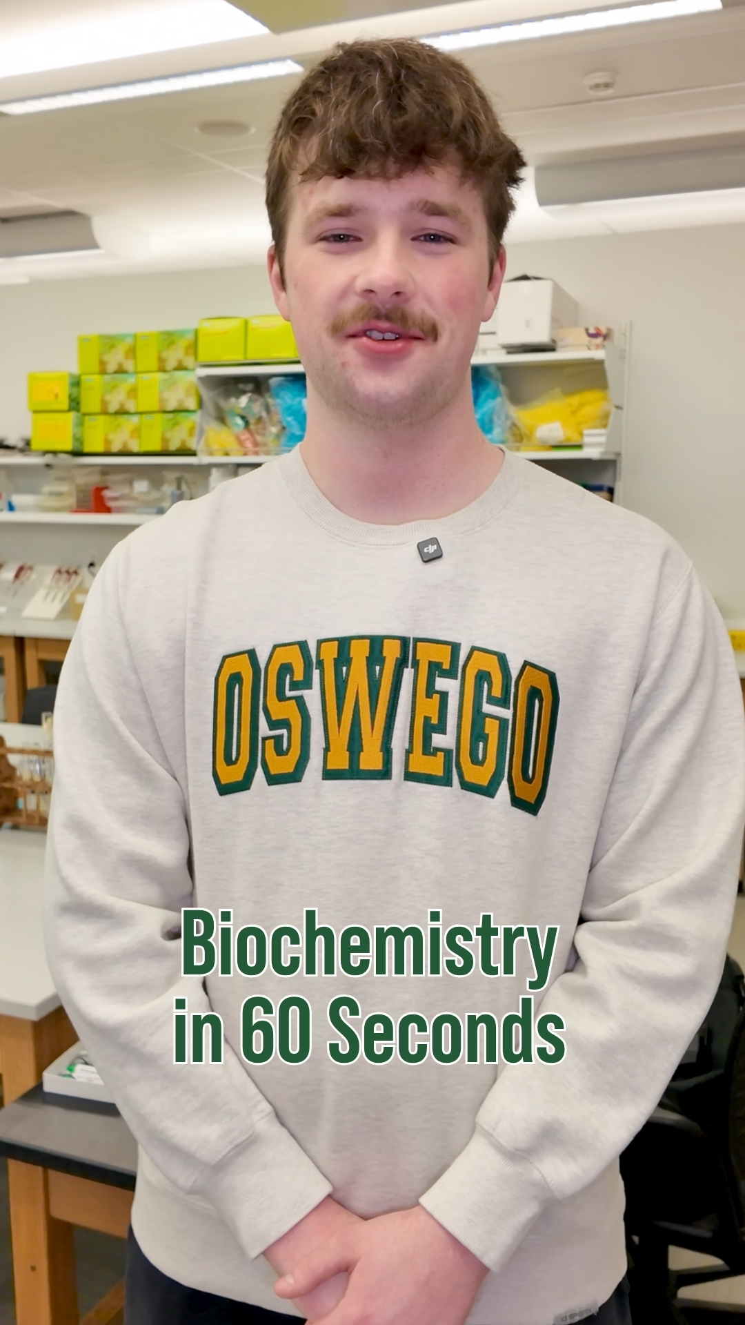 Student standing in a science lab wearing a gray Oswego sweatshirt. Lab supplies and equipment are visible in the background. On-screen text reads “Biochemistry in 60 Seconds.”