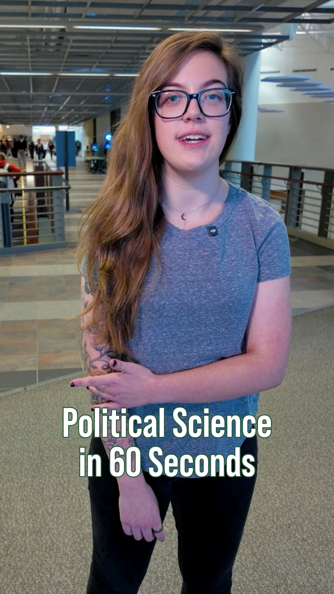 Student wearing glasses and a gray shirt standing inside a campus academic building with an open hallway and railing in the background. On-screen text reads “Political Science in 60 Seconds.”