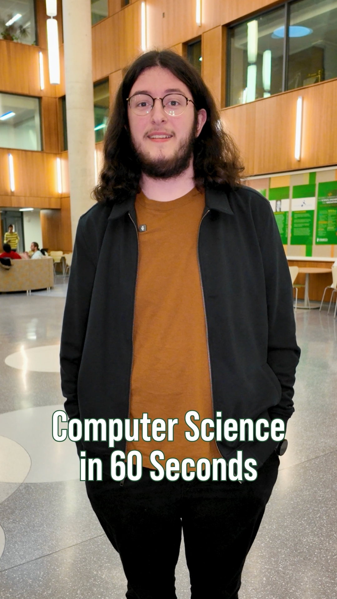 Student with long hair and glasses standing inside a modern campus academic building with wood-paneled walls and seating areas in the background. On-screen text reads “Computer Science in 60 Seconds.”