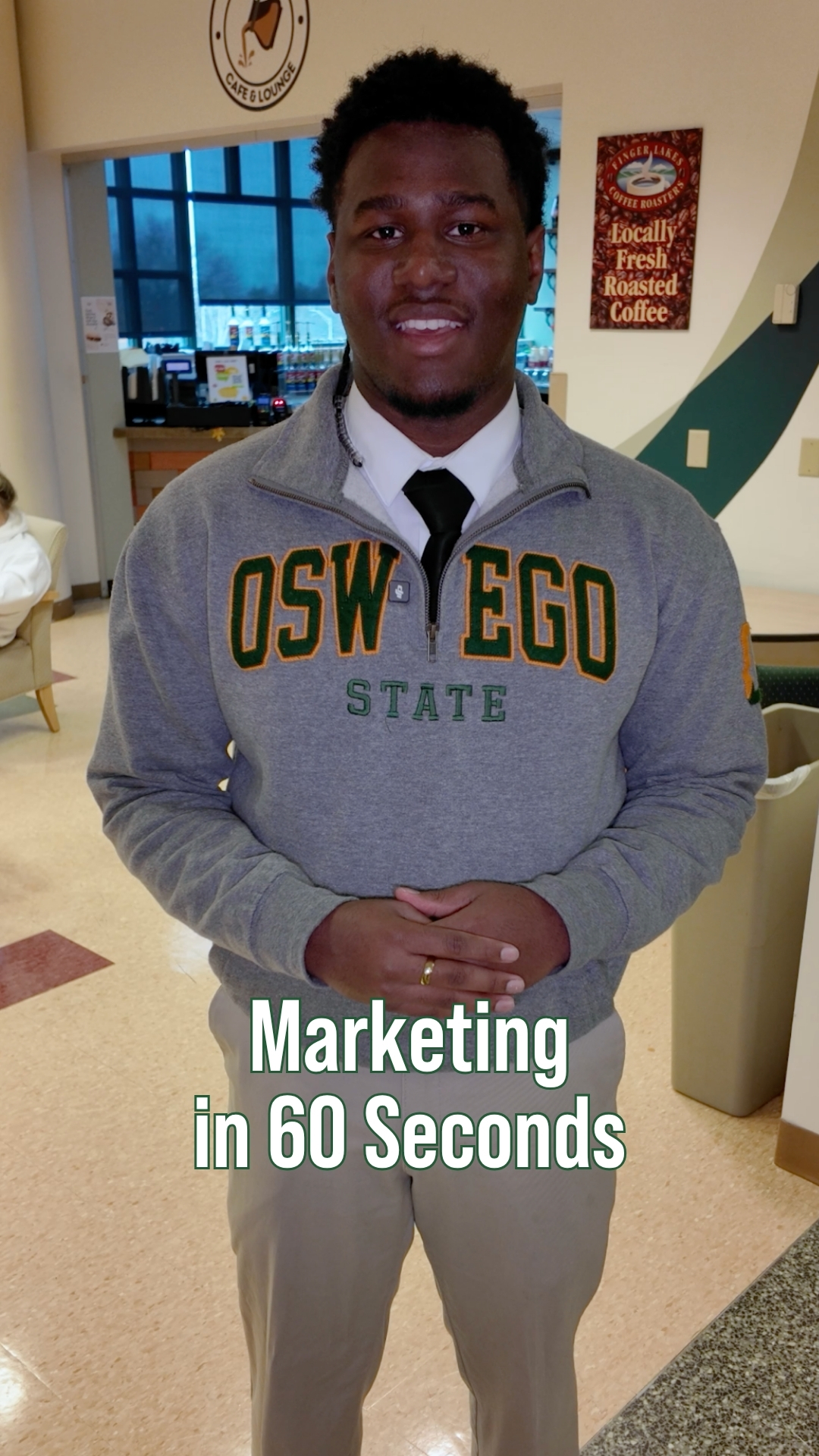 Student wearing a gray Oswego State quarter-zip and tie standing inside a campus café area. Coffee shop signs and seating are visible in the background. On-screen text reads “Marketing in 60 Seconds.”