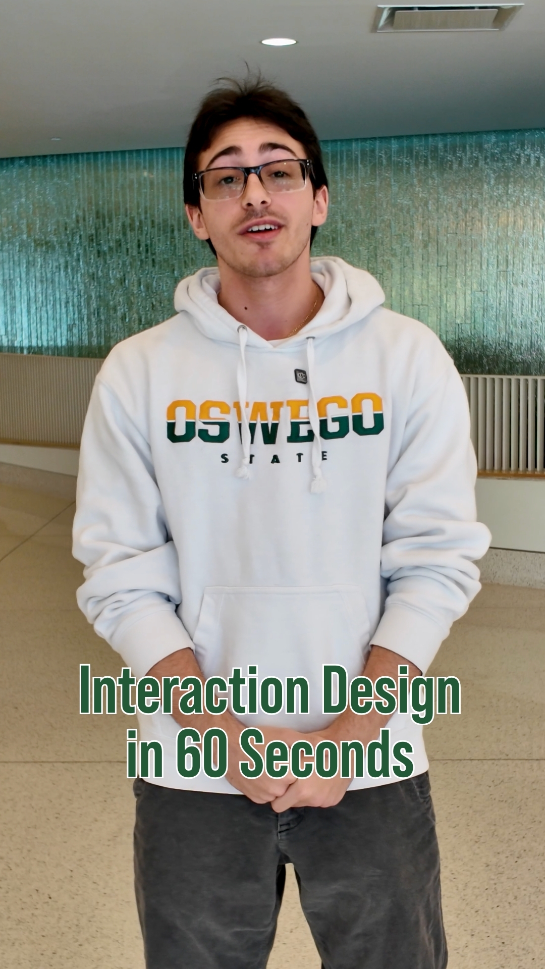Student wearing a white Oswego State hoodie and glasses standing inside a campus building. On-screen text reads “Interaction Design in 60 Seconds.”