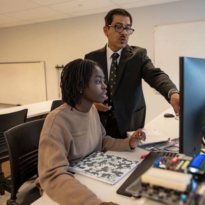 A professor stands beside a student in a classroom, pointing at a computer monitor while guiding them through work at a desk with electronics equipment and a notebook.