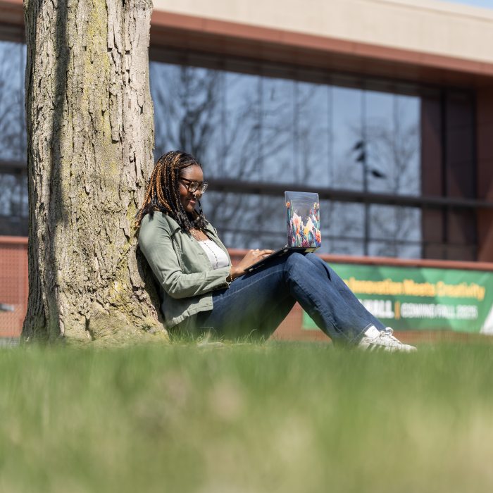 tudent working on a laptop while seated under a tree on campus.