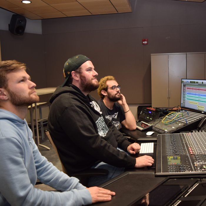 Three students in a professional recording studio, using a mixing console and digital audio workstation.