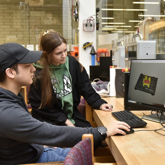 Two students use 3D modeling software at a computer workstation in a technology education lab.