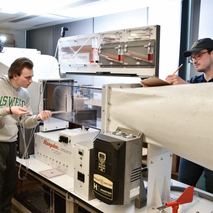 Two meteorology students collect data from a wind tunnel experiment in a laboratory setting.