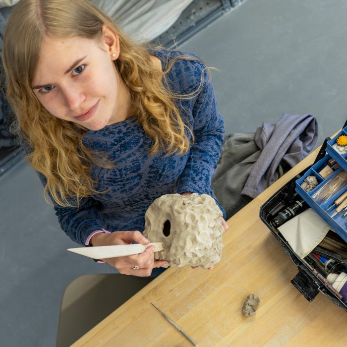 A studio art student uses sculpting tools to shape a ceramic artwork at a worktable.