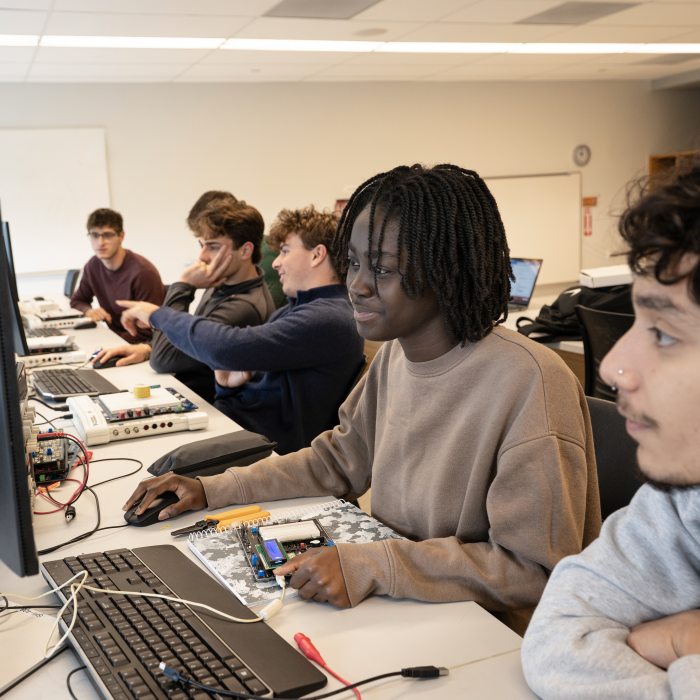 Students in a classroom, looking at computer monitors at a desk with electronics equipment.