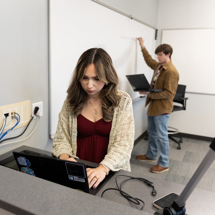 Student looking at a laptop while another writes on a whiteboard in the background.