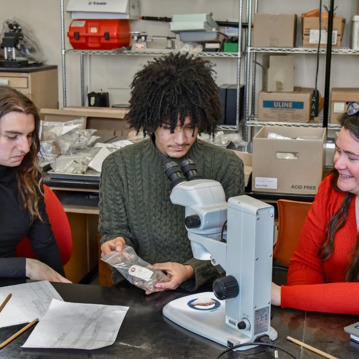 An anthropology professor and two students examine research specimens using laboratory equipment.