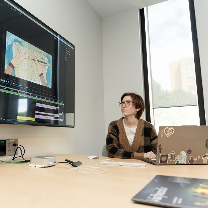 A student works at a laptop while viewing digital media editing software on a large display screen.
