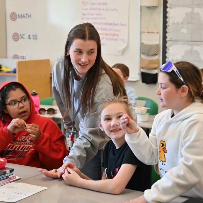 Undergraduate student teacher with three students around their desks looking at stones.