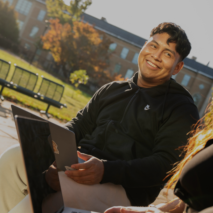 Student sits outdoors with a laptop, smiling while talking with another student on a sunny campus walkway, as two students work on laptops in the background.