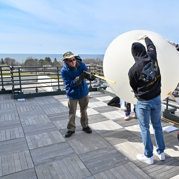 Students and a faculty member inflate a large white weather balloon on a rooftop overlooking the water