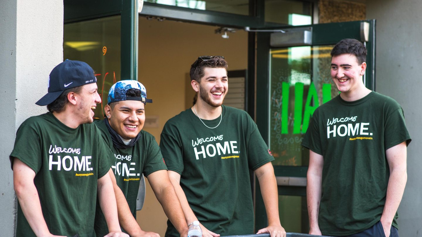 Four male students laughing and wearing green &quot;Welcome Home&quot; T-shirts stand around moving bins