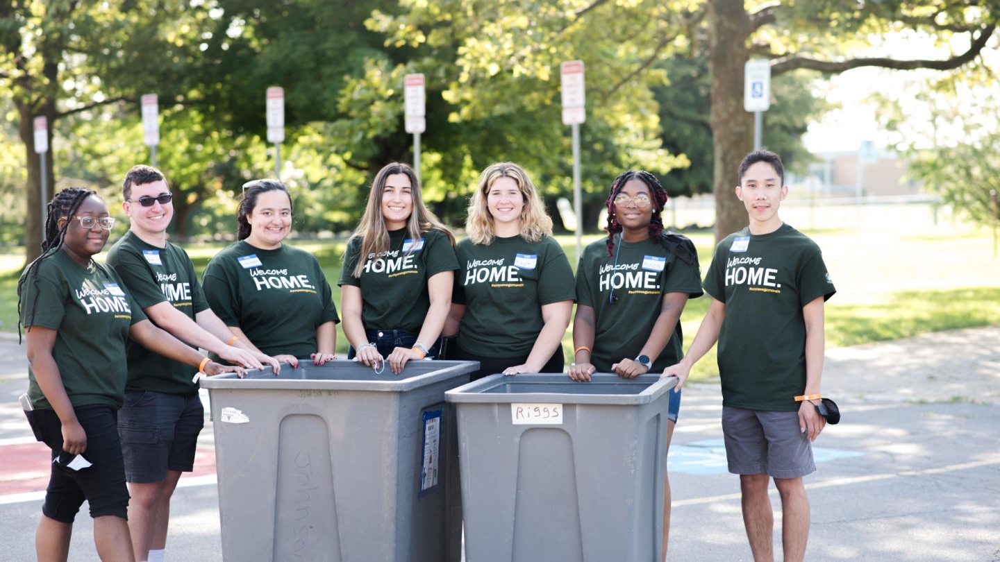 A group of students wearing green &quot;Welcome Home&quot; t-shirts pose by two moving bins