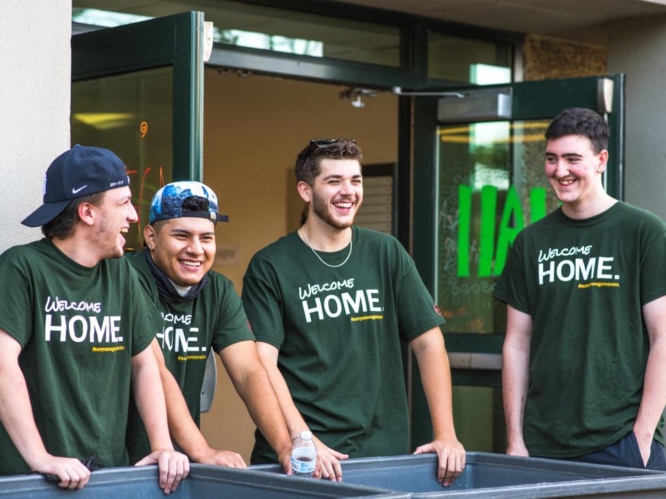 Four male students laughing and wearing green &quot;Welcome Home&quot; T-shirts stand around moving bins