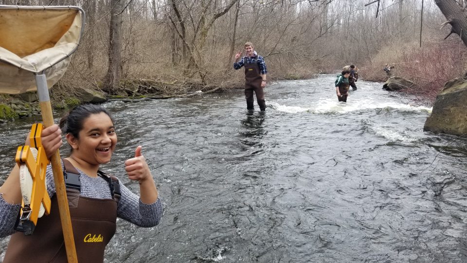 Three people stand in a stream wearing chest waders and measuring tape