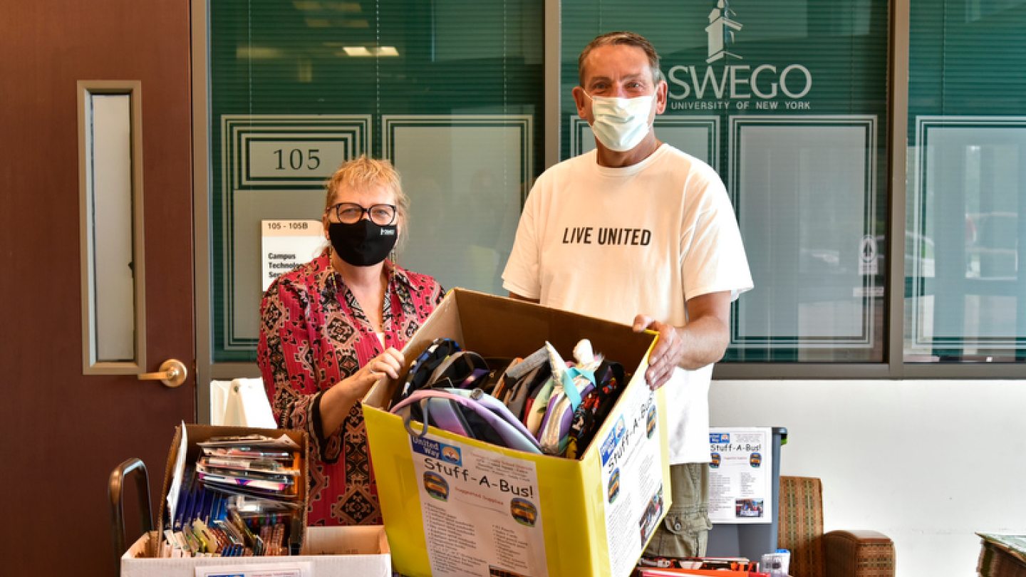 Two people wearing masks, standing behind boxes labeled &#039;Stuff-A-Bus&#039; filled with donated school supplies, in front of a door and window with a SUNY Oswego logo.