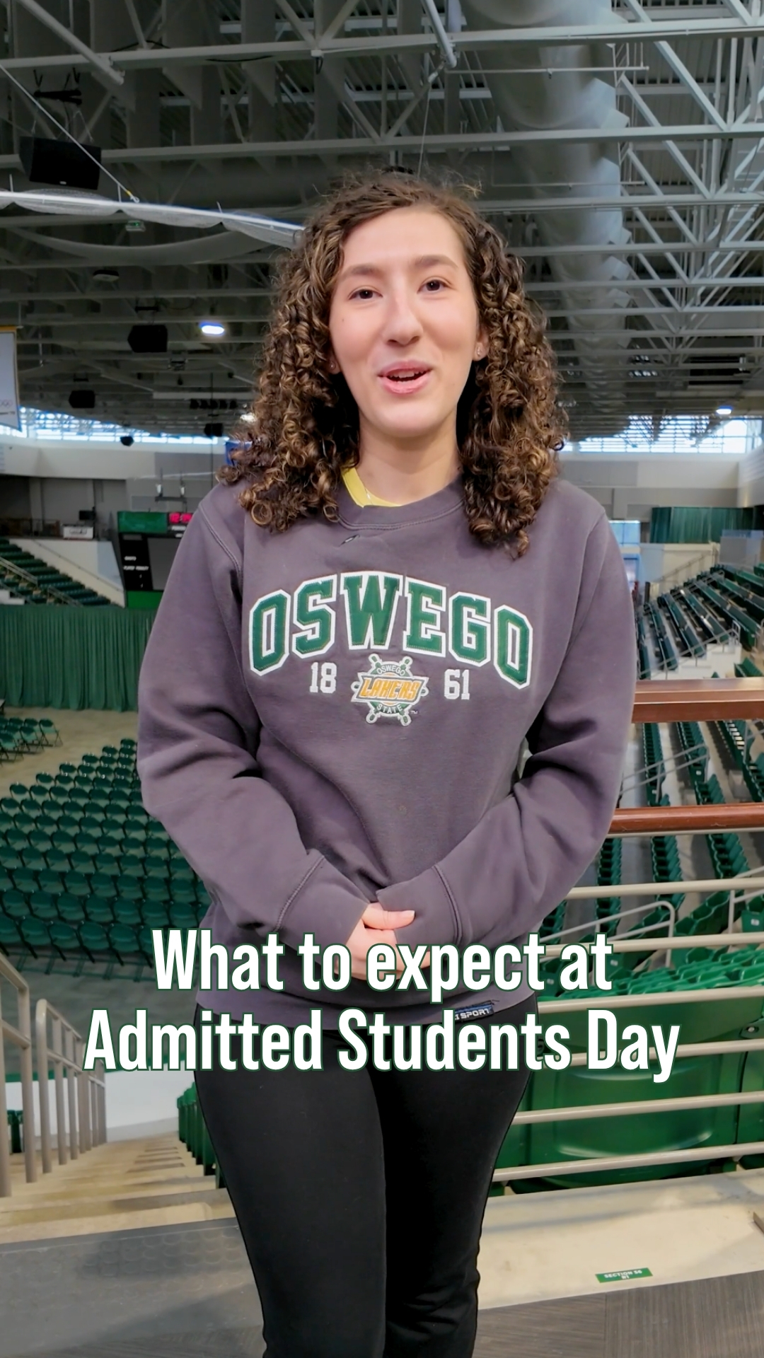 A student wearing an Oswego sweatshirt stands in a large indoor arena with rows of green seats, speaking to camera with text on screen reading “What to expect at Admitted Students Day.”