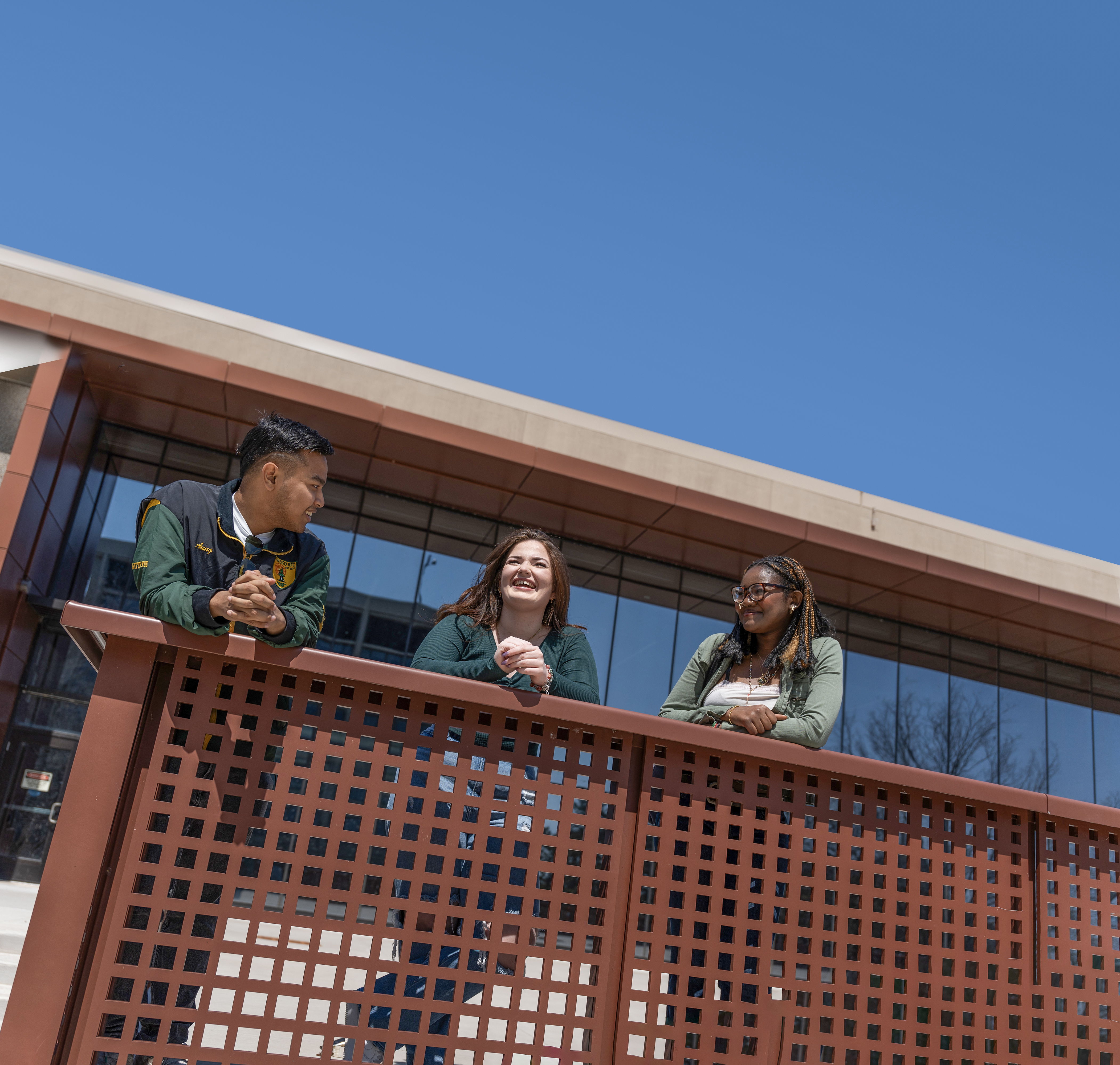Three students stand along a railing outside a modern building under a clear blue sky.
