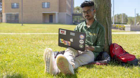 A student sits under a tree with his laptop.