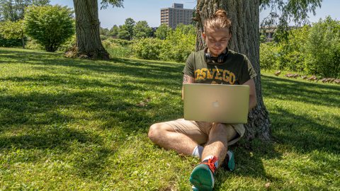 A student studies on a laptop under a tree next to glimmerglass lagoon