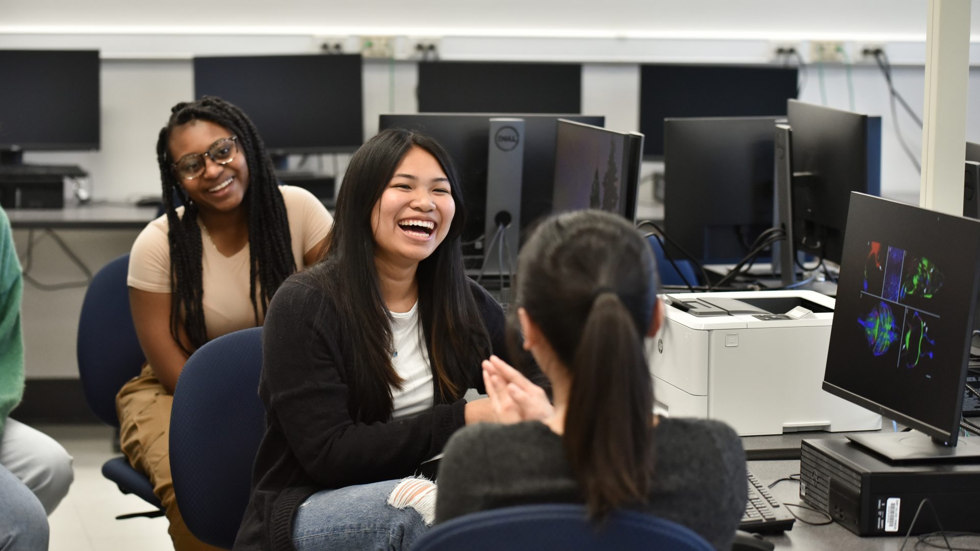 Several students sit together in a computer lab, smiling and talking during class while desktop monitors and colorful images displayed on a screen are visible in the background.