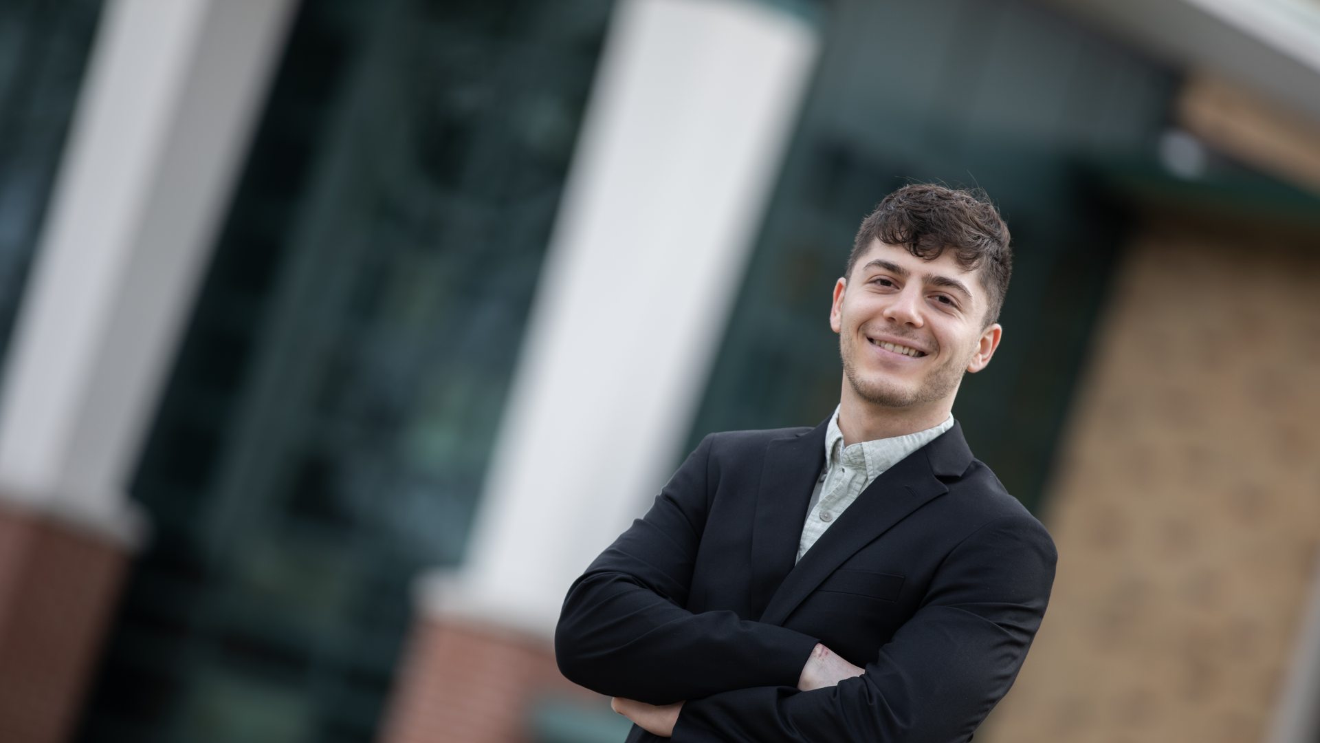 A smiling student wearing a dark blazer and light button-down shirt stands with arms crossed outside a campus building, posing for a professional portrait with a blurred background.