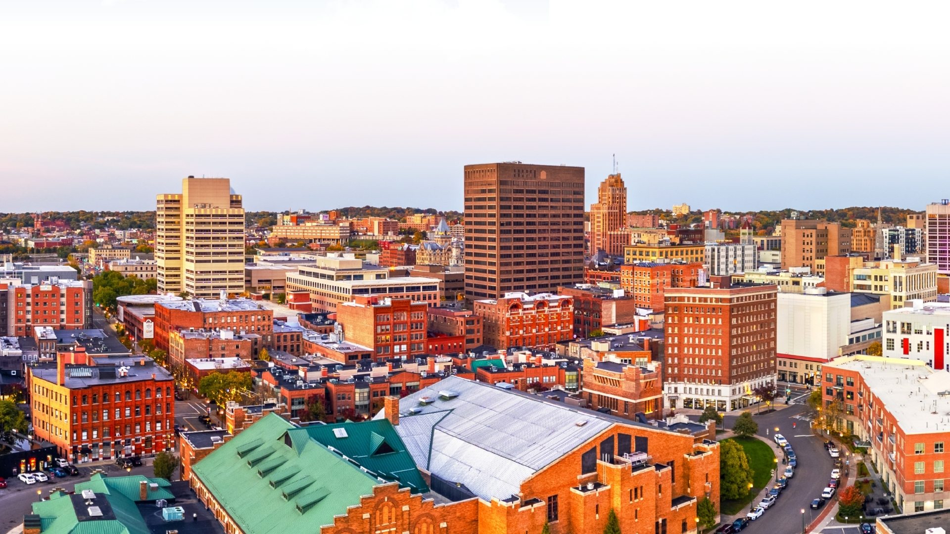 A panoramic view of downtown Syracuse, New York, featuring a mix of historic brick buildings, modern office towers and city streets at sunset under a clear evening sky.