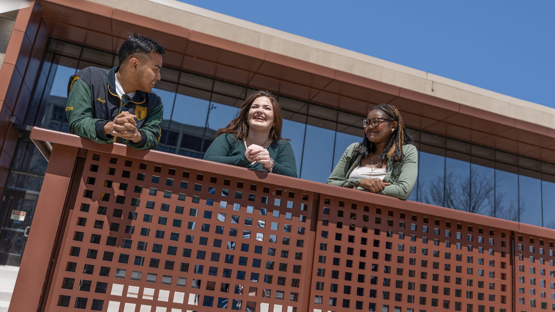 Three students stand along a railing outside a modern building under a clear blue sky.