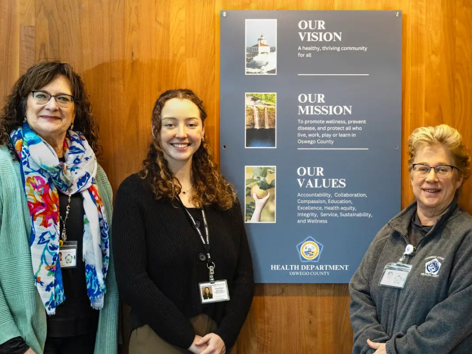 Tessa Mucha standing with two colleagues at the Oswego County Health Department in front of a mission and values display.