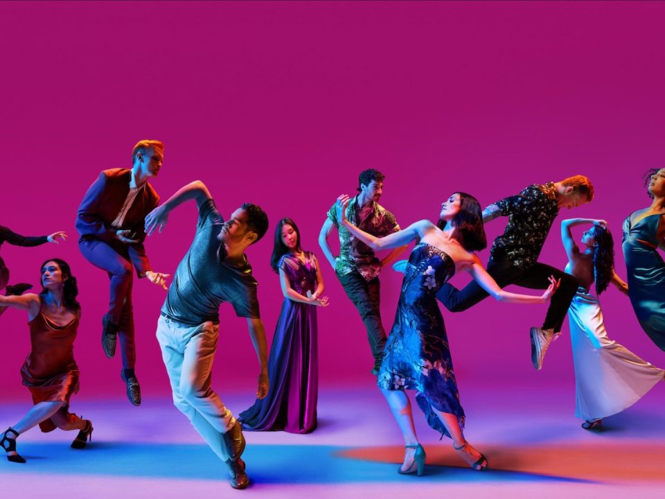 A group of dancers in blue costumes performing on a stage lit in pink