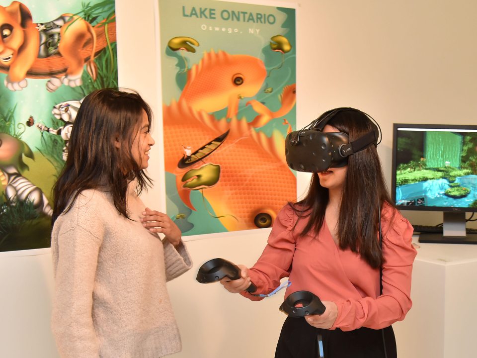 Two students in front of colorful artwork, one wearing a VR headset and holding controllers while the other smiles.