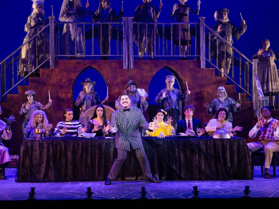 Cast members of The Addams Family sitting in costume at a long table, with ensemble members on stairs behind them.