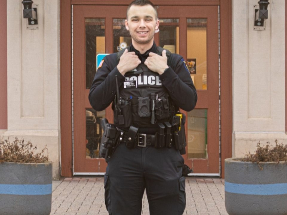 Officer Gombas stands in uniform in front of the entrance to the Oswego City Police station.