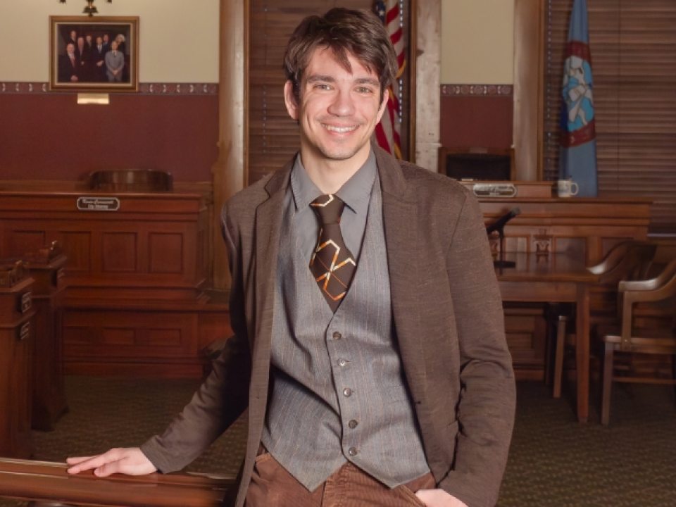 Greg Caster, wearing a suit jacket, smiles inside Oswego’s historic City Hall, with dark wood trim, a brass chandelier, and the City of Oswego flag in the background.