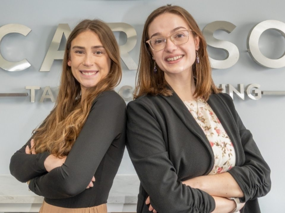 Alexis Dardaris and Kymberly Terwilliger standing back to back with arms crossed in front of “Caruso Tax Accounting” sign.
