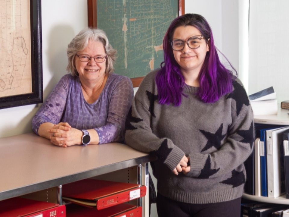 Debra Allen (left) and Madison Getwright (right) stand next to a shelf of red archive boxes surrounded by framed historic parcel maps. 