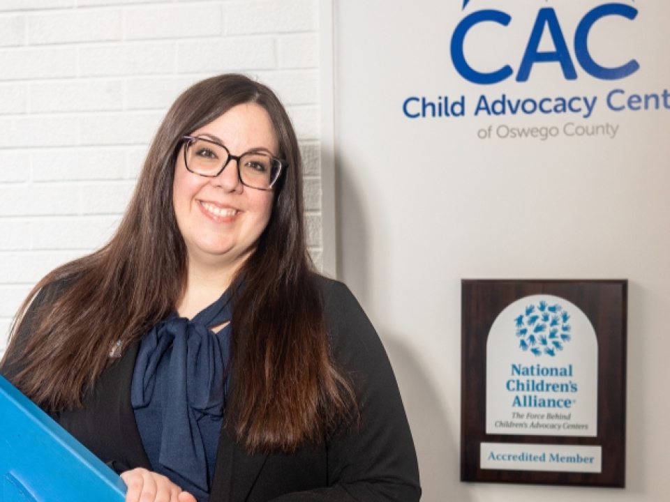 Melanie Smith stands on a blue staircase, smiling at the camera. She is wearing a dark jacket over a blue top. Behind her, on the wall, is a sign for the Child Advocacy Center of Oswego County