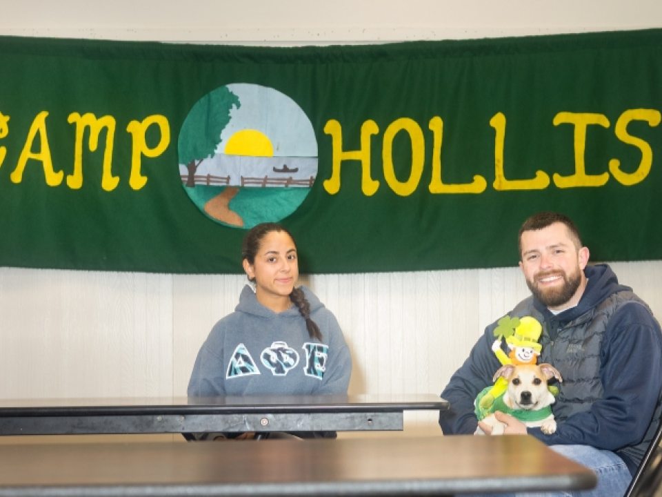 Izzy Ronacher (left) and Kyle Perez (right) seated at a table with a banner that reads "CAMP HOLLIS" in the background.