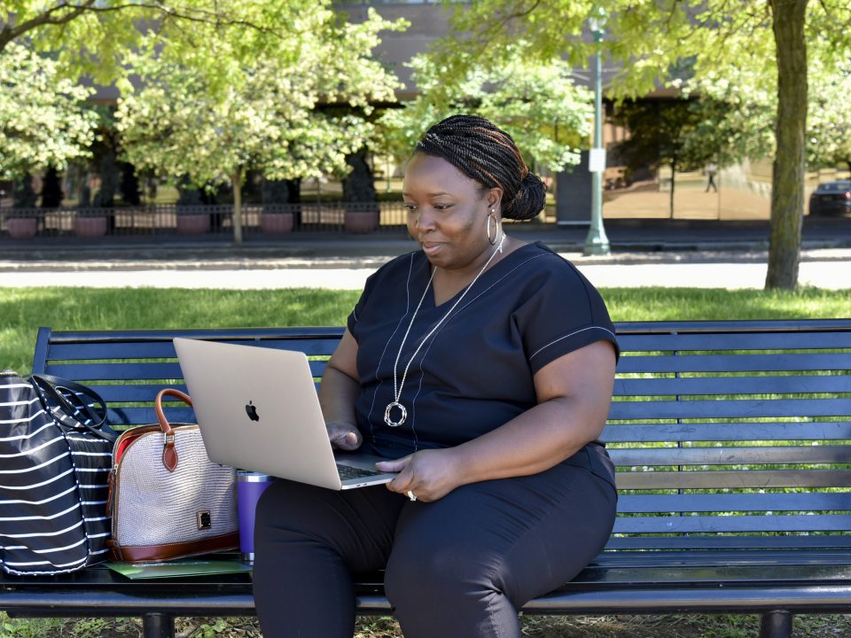 An adult student works on a laptop while sitting on a bench outside in downtown Syracuse, balancing coursework with a busy lifestyle.
