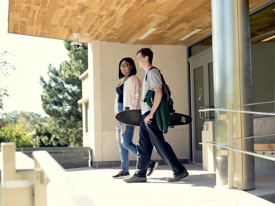 Two students walk together outside a campus residence hall, smiling as one carries a skateboard and the other wears a backpack.