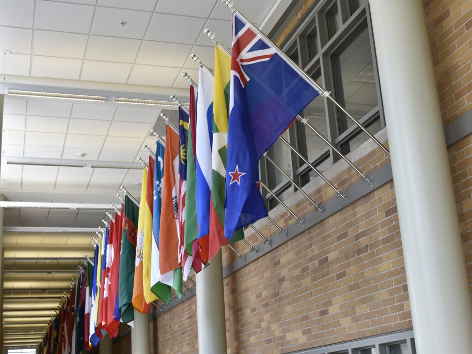 International flags hang along a bright campus hallway, representing countries from around the world.