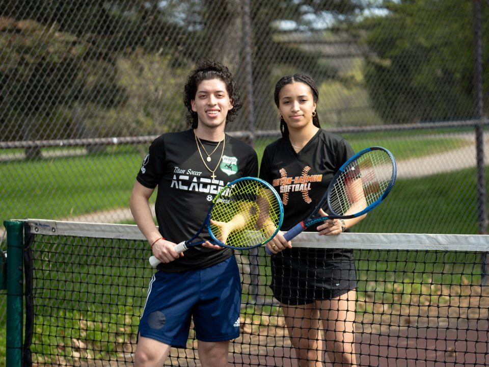 Two students holding tennis rackets stand together on an outdoor tennis court.